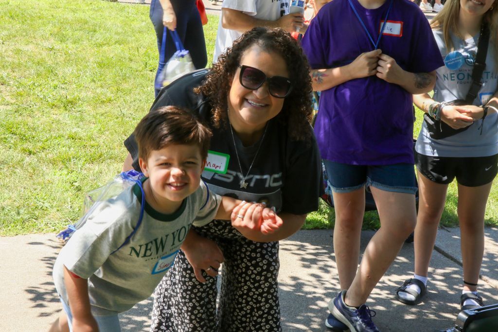 Woman and child smiling, outside. Woman holds child's hand. Others in background.