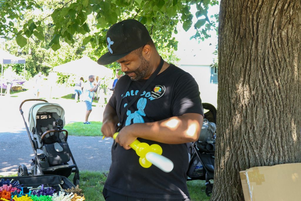 Man in black shirt and cap making a balloon sword at an outdoor event, next to a tree and strollers.