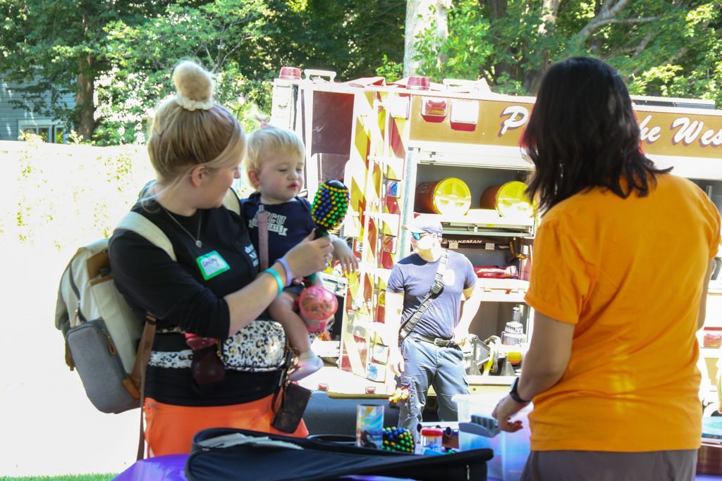 Woman holding child, showing a maraca. Event with fire truck and person in orange shirt.