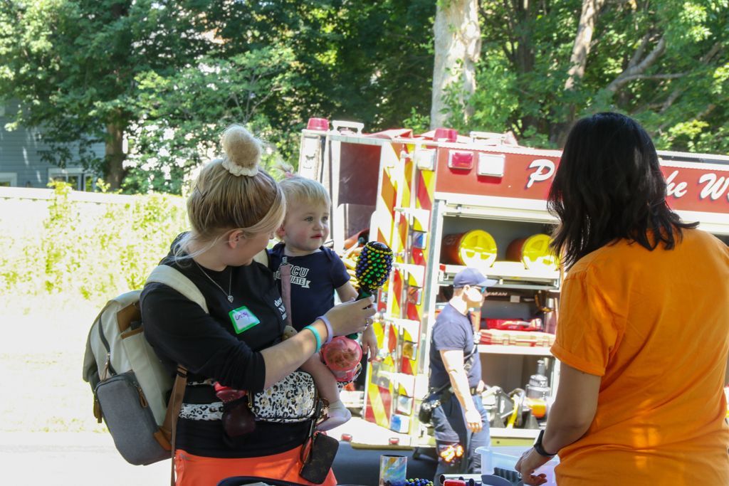 Woman holding a child points toward a fire truck at an outdoor event, another person watches nearby.