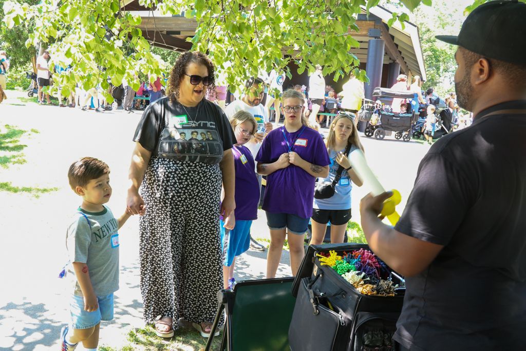Group of people watching a balloon artist at an outdoor event, some children looking on.