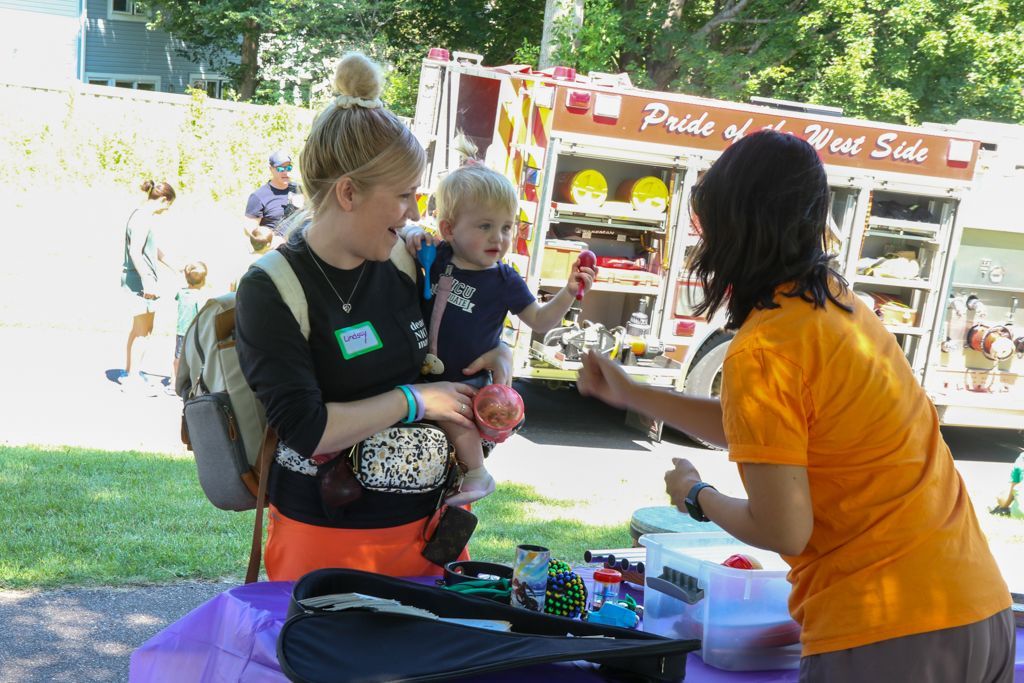 Woman holds child near table with toys, interacting with another person in front of a fire truck.