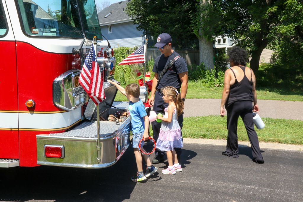 Firefighter showing kids fire truck at an outdoor event. Red and white truck, American flag, children, adult.