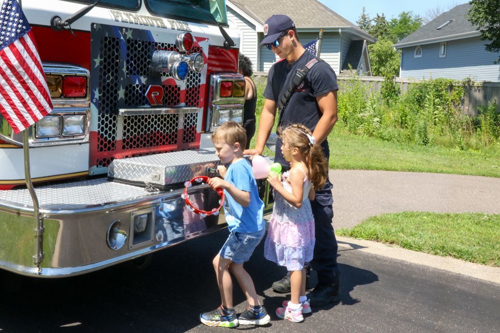 Firefighter helps two children explore a fire truck. American flag, bright sunny day.
