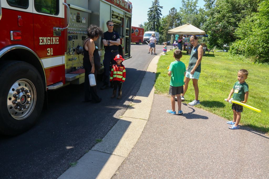 Fire truck at community event; child in firefighter outfit, people on sidewalk, sunny day.