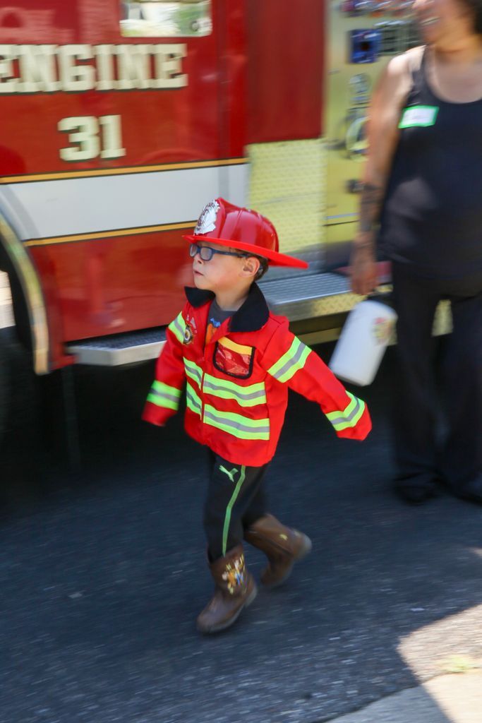 Young child in a firefighter costume walks past a fire truck.
