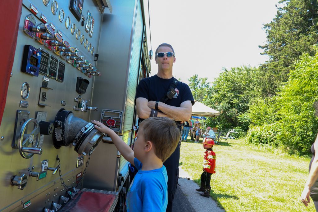 Boy points at fire truck controls as firefighter watches. Outdoors, sunny day.