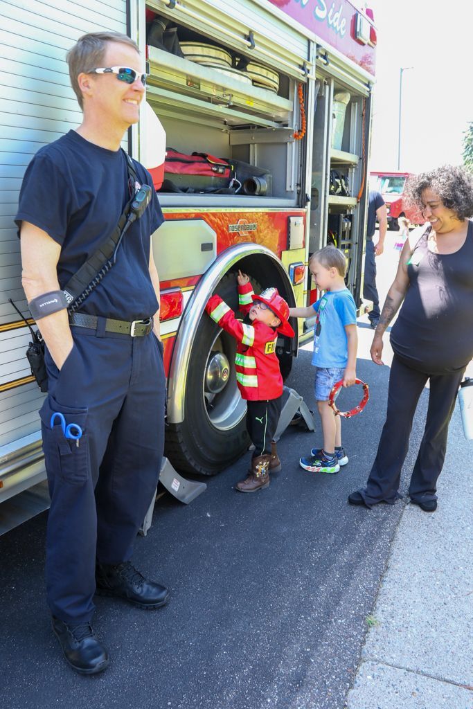 Firefighter stands near fire truck as children explore it and play with a woman.