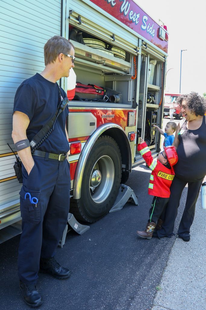 Firefighter stands near fire truck while a child in a costume points at the truck with a woman watching.