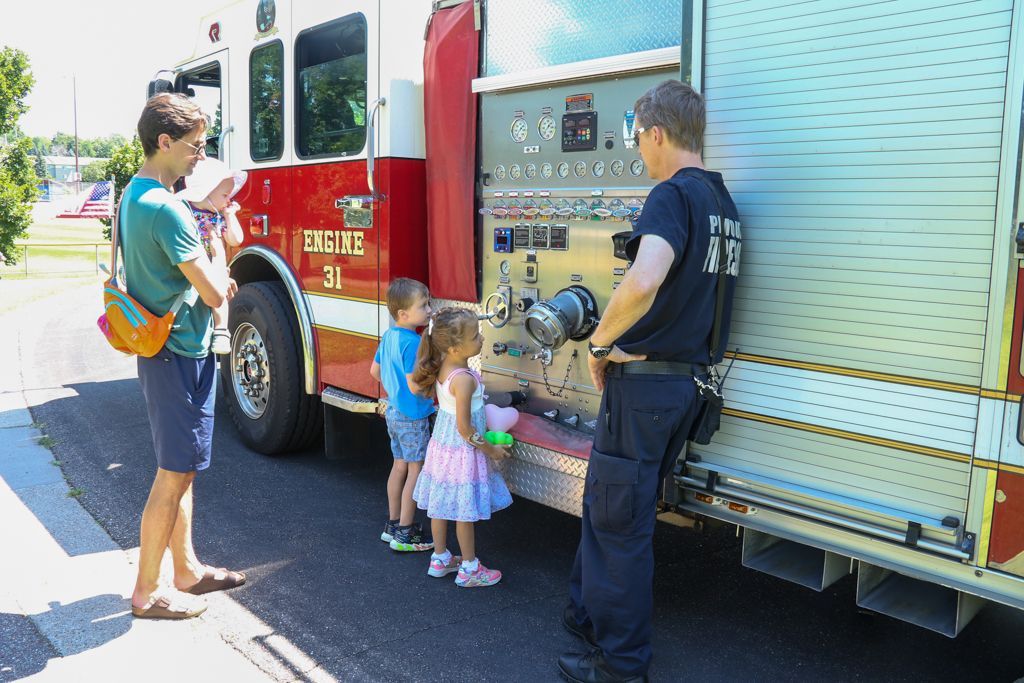 Firefighter showing children the controls on a fire truck; an adult looks on.