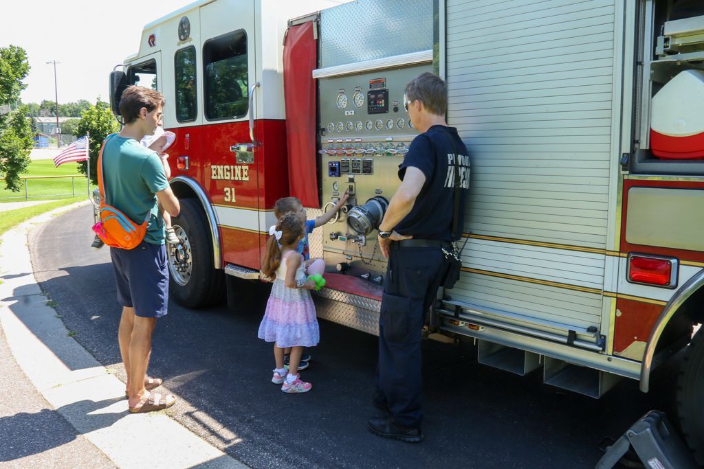 Firefighter shows a young child and adult fire truck controls outside. Red and white truck, sunny day.