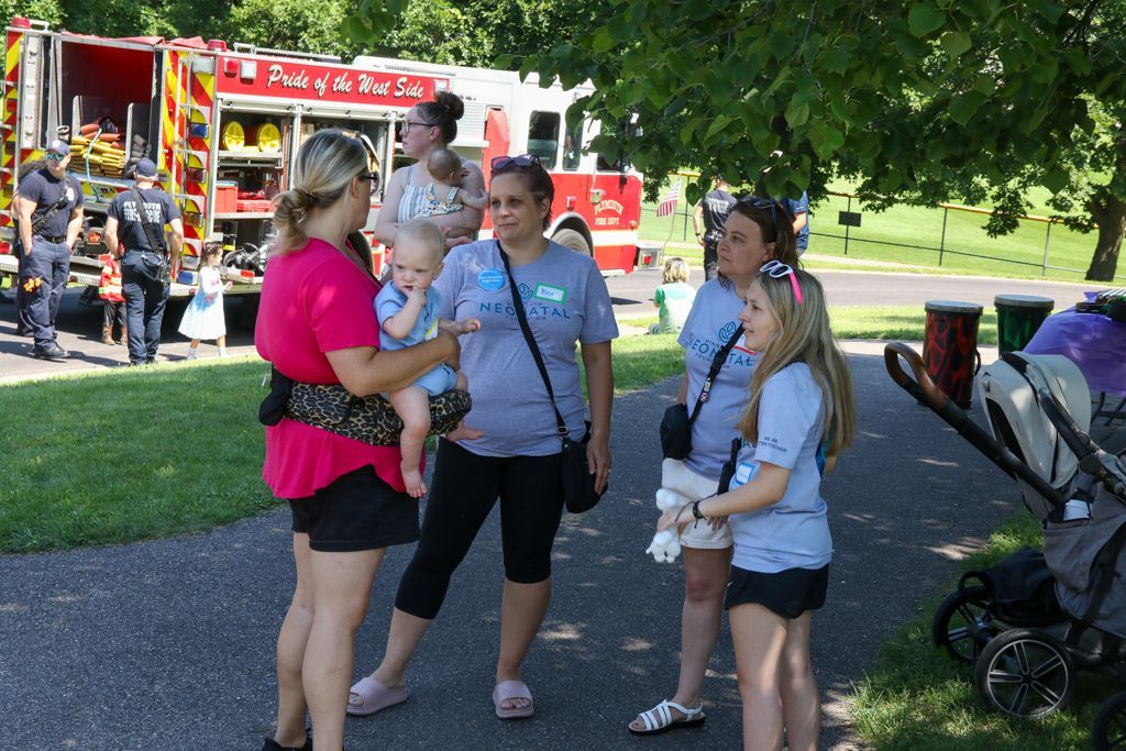 People in front of a fire truck. Two women hold babies, others chat on a path in a park.