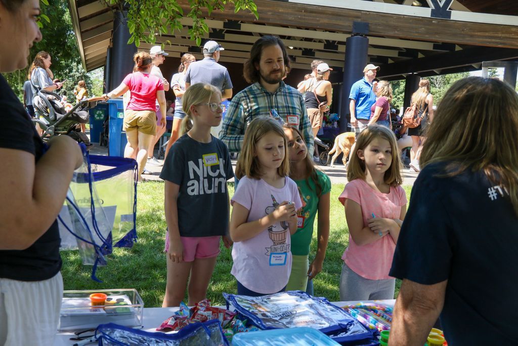 Children at a table with adults, outdoor event. People in the background. Sunny day.
