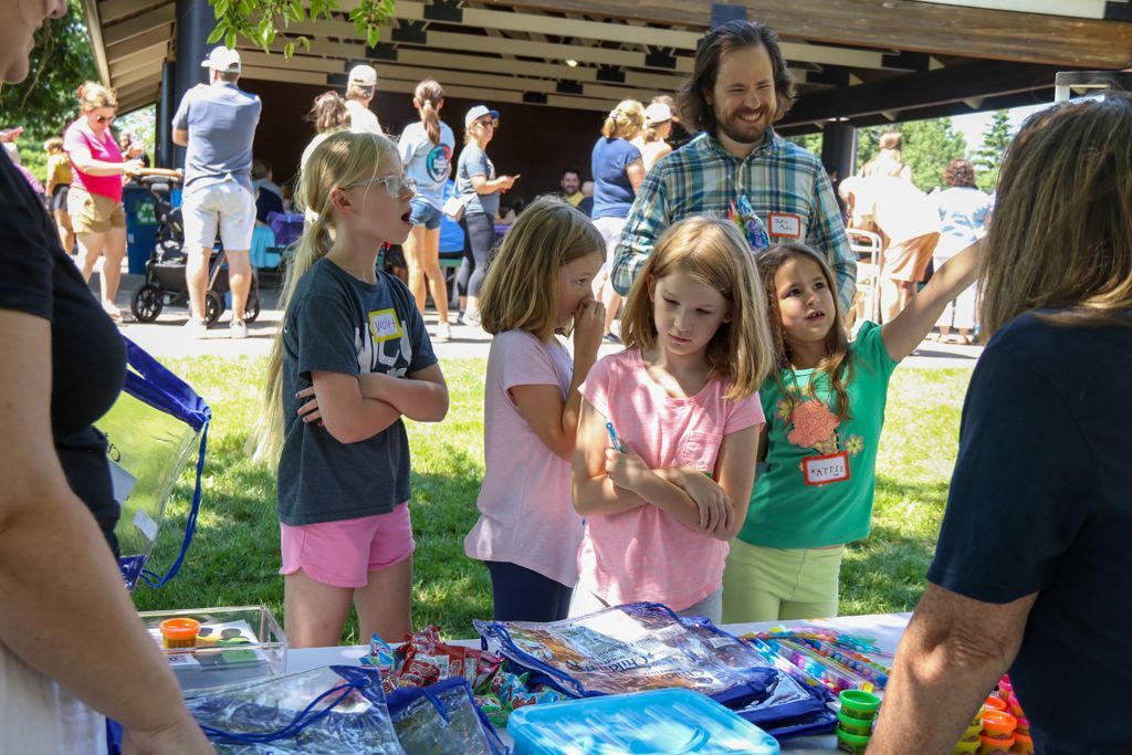 Children looking at a craft table outdoors with an adult, other people in the background.