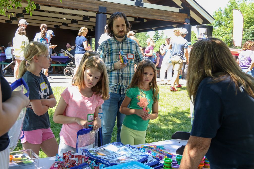 Children look at items on a table at an outdoor event, attended by others in a park setting.