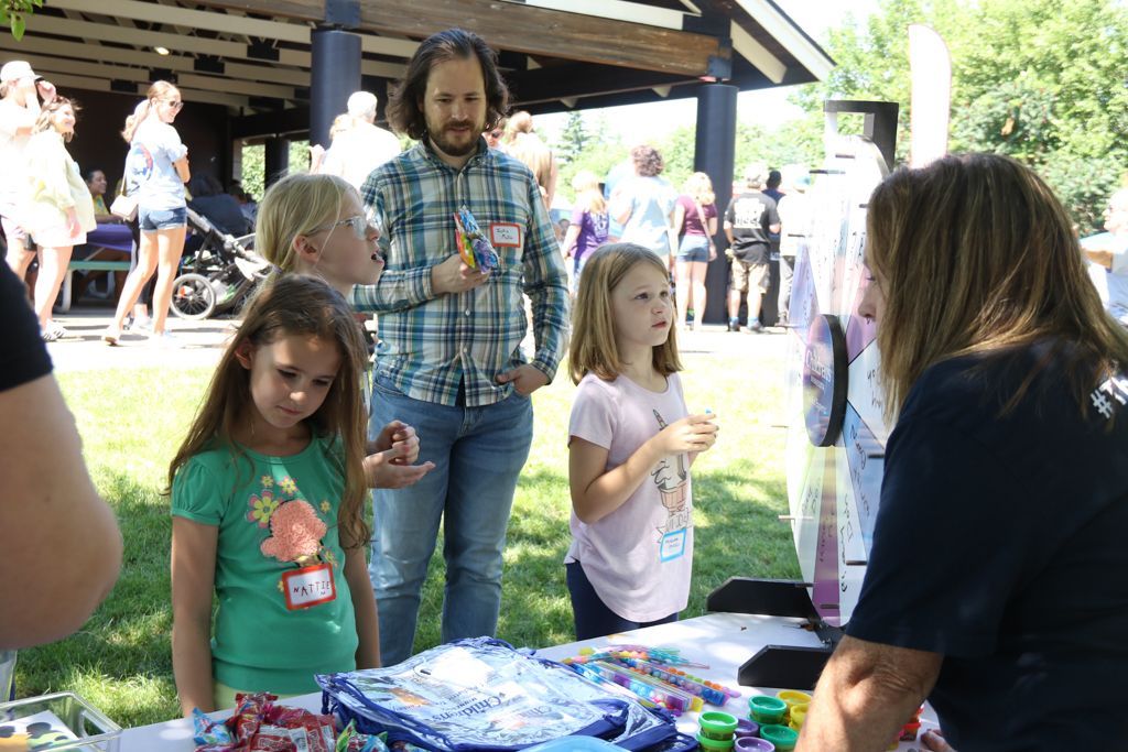 People at a table. Three children with an adult browse items for sale at an outdoor event.