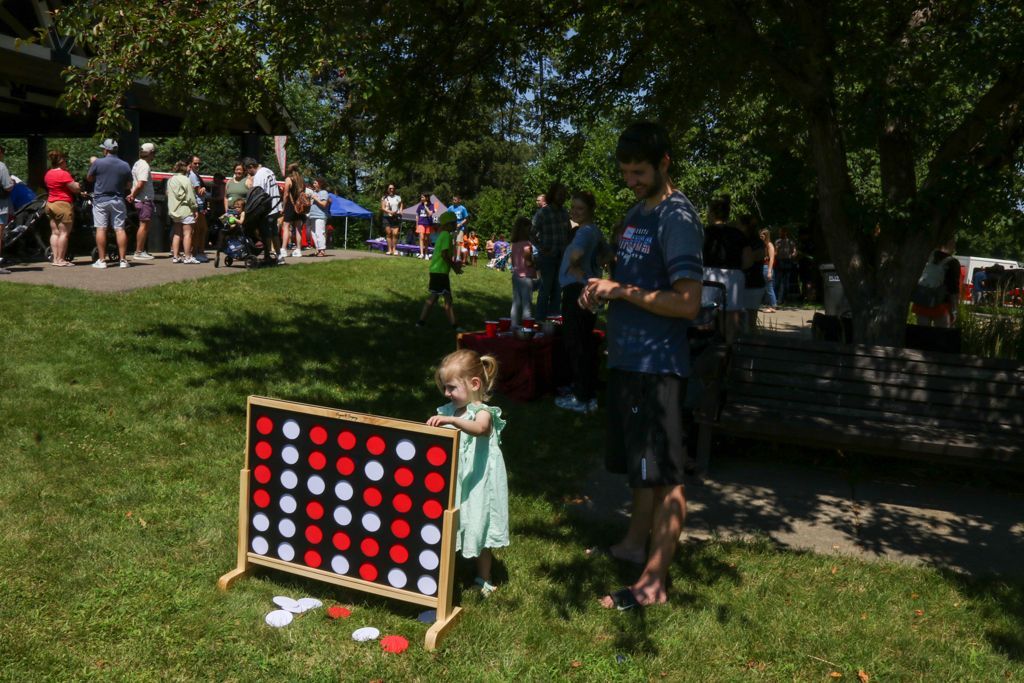 People playing giant Connect Four in a park on a sunny day. A child and man stand nearby.