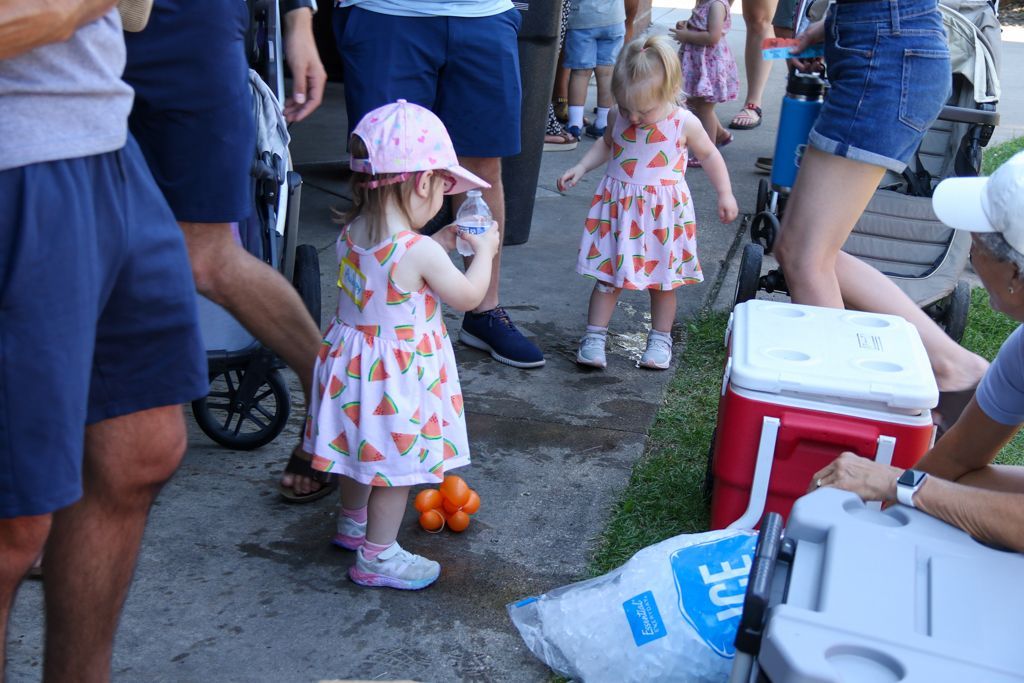 Two young children at a gathering, one in a pink hat, playing with oranges near coolers, surrounded by people.