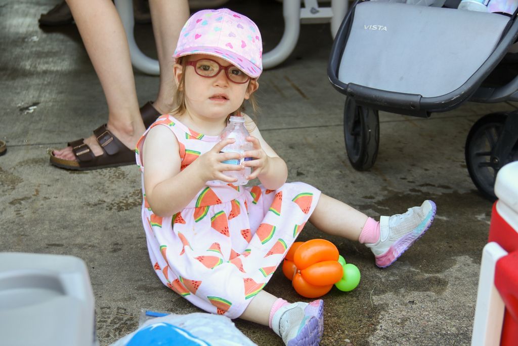 Child wearing glasses and watermelon dress, sitting on the ground, holding a water bottle.