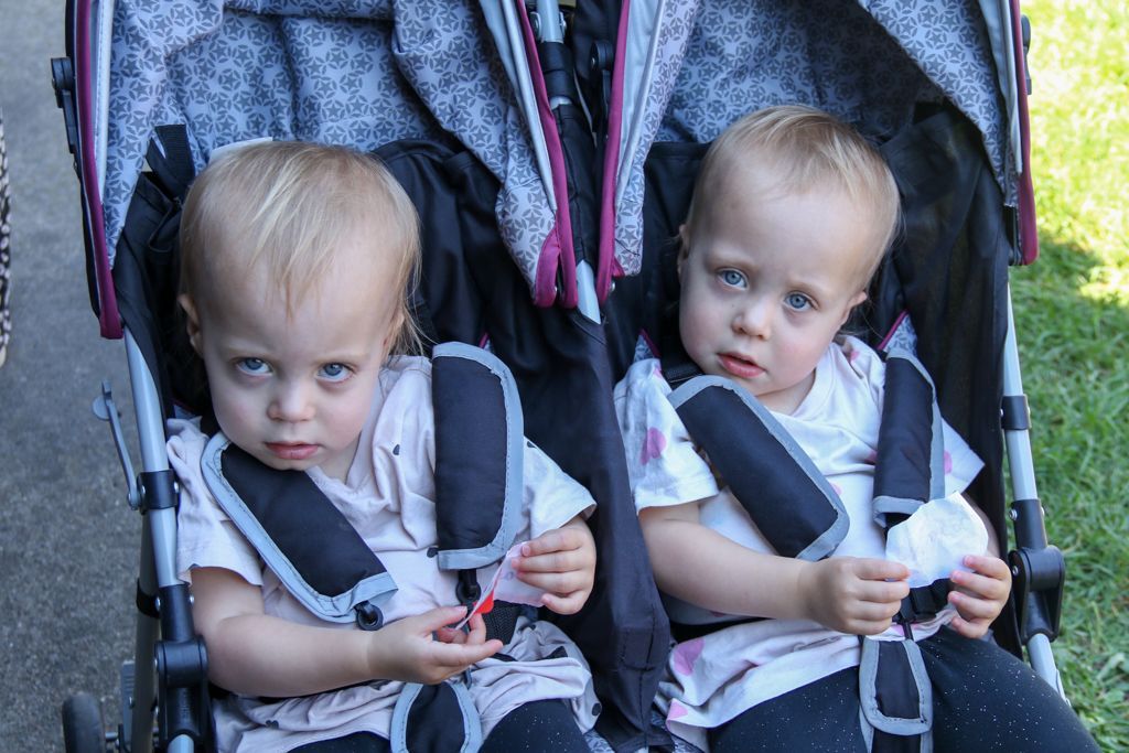 Two fair-haired toddlers with blue eyes sit in a double stroller, looking forward with serious expressions.