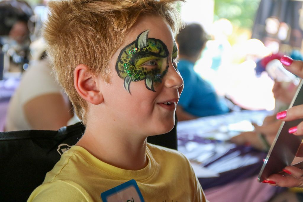 Boy with face painted like a dragon eye, smiling, outdoors.