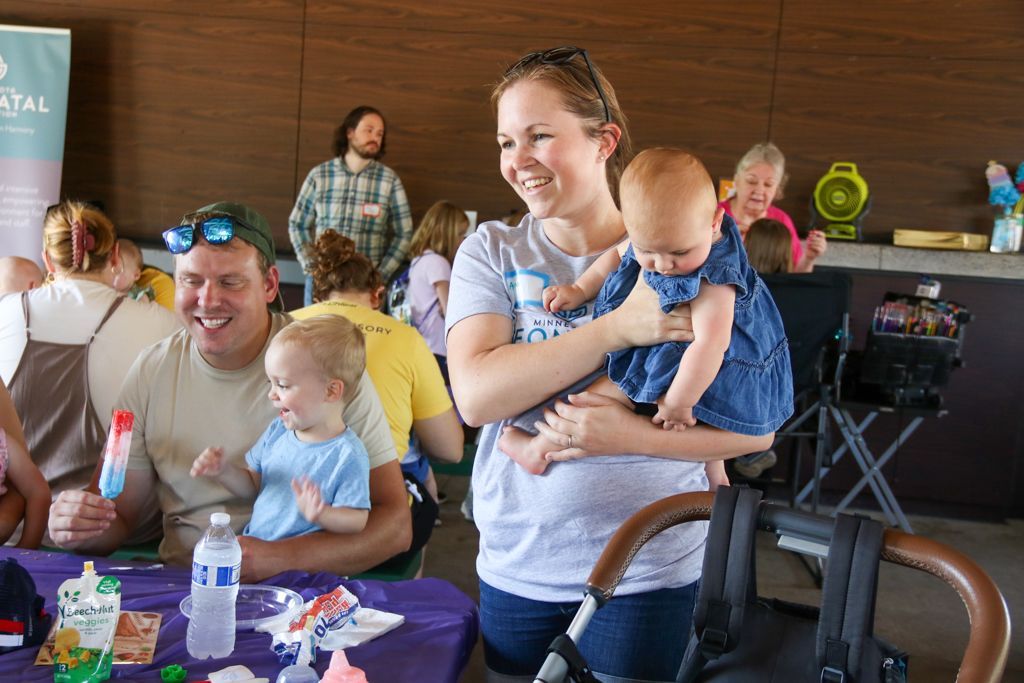 Family at an outdoor event; parents hold children, smiling. Purple table with snacks, balloons in background.