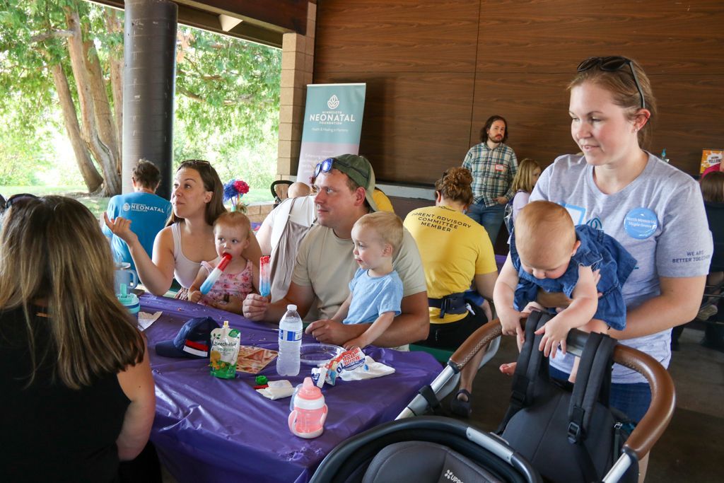 People gathered at an outdoor event with babies. Several adults at a table, some holding infants. Purple tablecloth, trees in background.