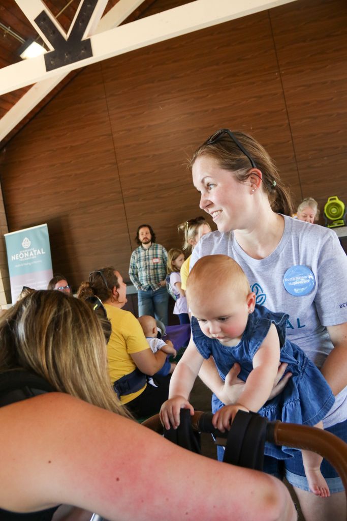 Woman holds a baby, looking at a stroller, inside a brown building with other people.