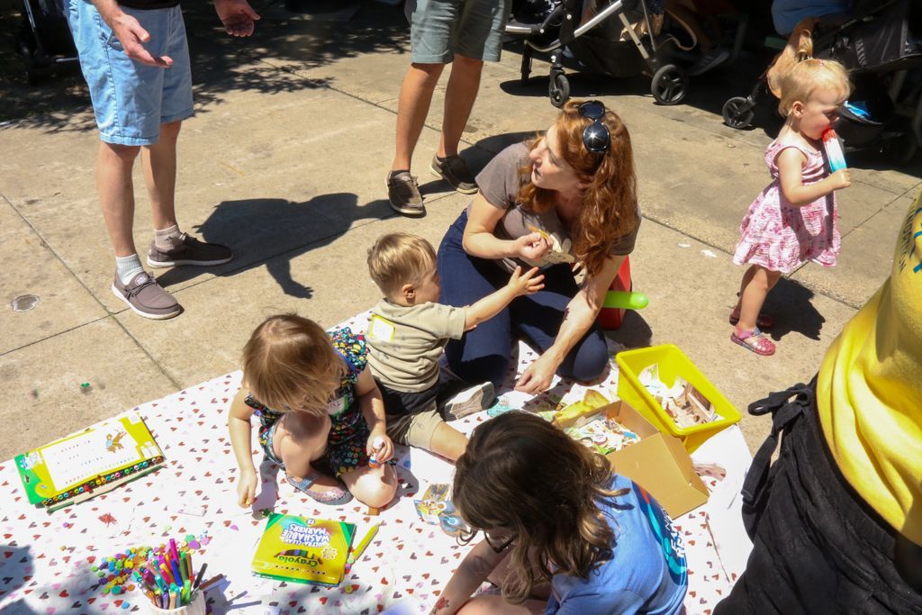 People on a sidewalk engaged in a picnic-style activity, including art, with children and adults.