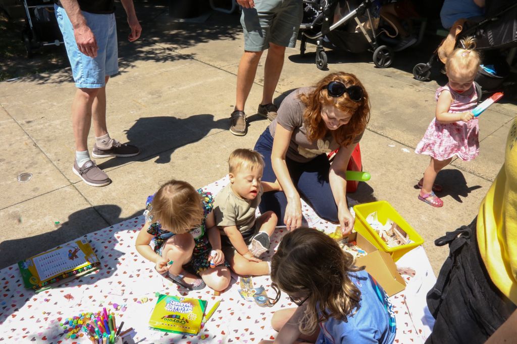 Woman and children sit on a blanket, drawing. A child holds a water gun. Outdoors in daylight.