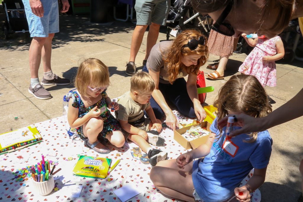 Children and a woman at a table, drawing. Outdoors, sunny.