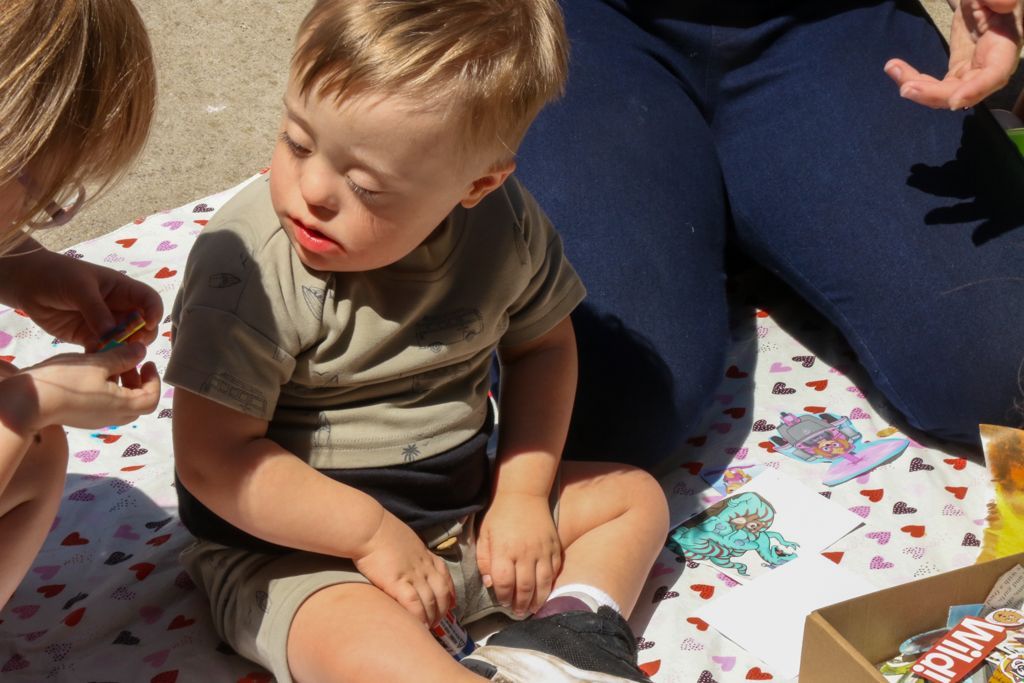 Boy with Down syndrome sitting on a blanket, looking down. Another person and legs are visible.