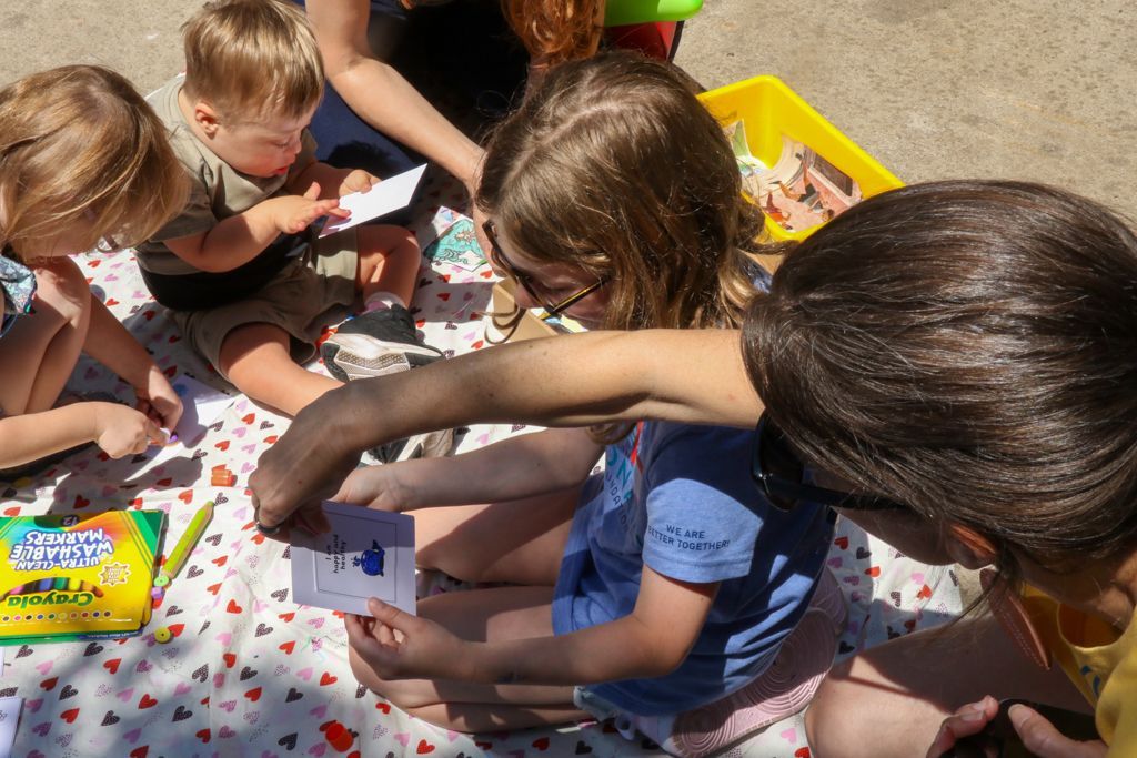 Children and adults gather around, looking at cards on a blanket outdoors.