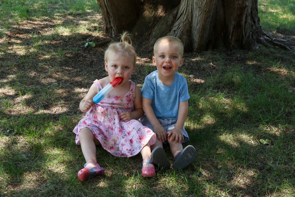 Two young children sitting on grass near a tree, one eating a popsicle, both smiling.