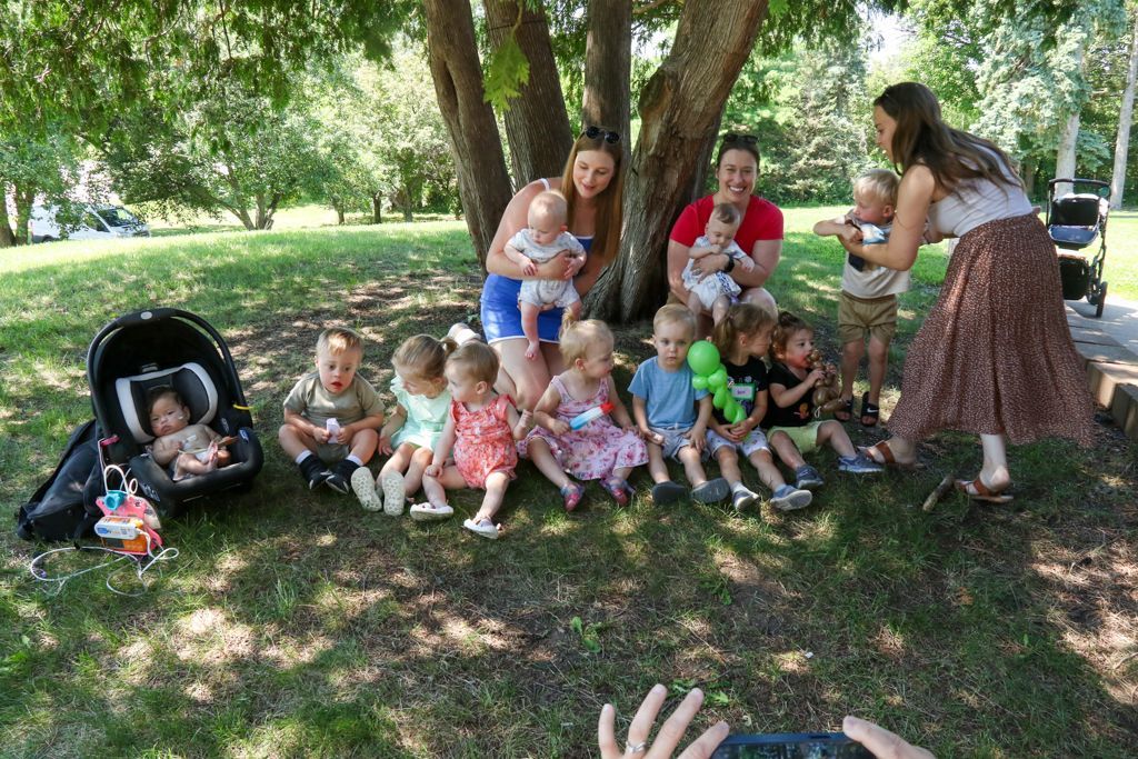 Group of toddlers and caregivers posing under a tree in a park; some sitting, some held; taking photo.