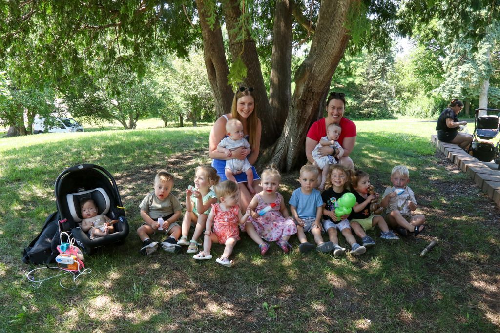 Group of babies and women sitting under a tree, outdoors. One baby in a car seat.
