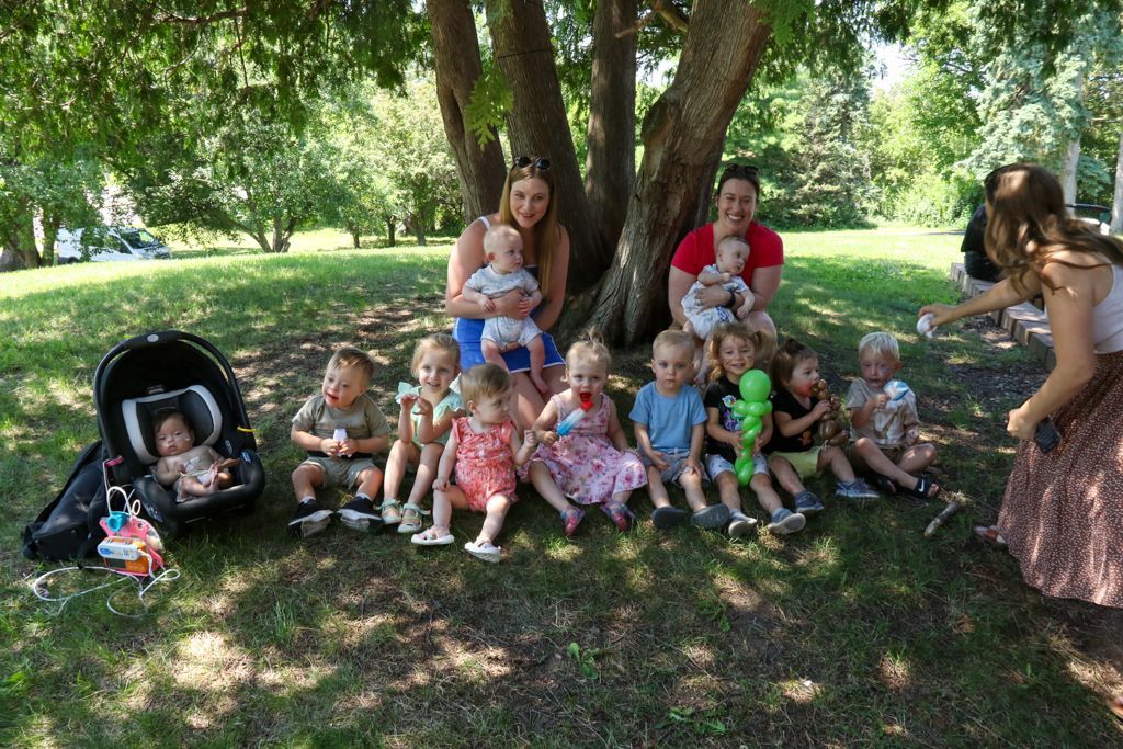 Group of babies and adults under a tree in a park. Babies sit on the ground, adults hold babies.