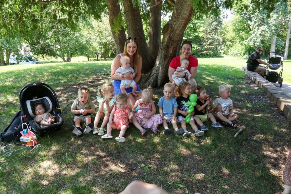 Group of babies and women posing for a photo under a tree in a park.