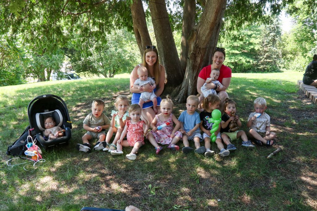 Group of children and two adults sitting under a tree on a sunny day.
