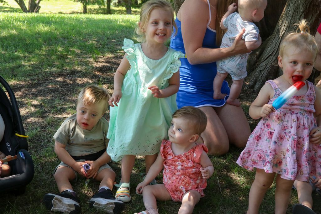 Children in a park, some sitting, some standing, one eating a popsicle. A woman holds a baby. Green grass, sunny day.