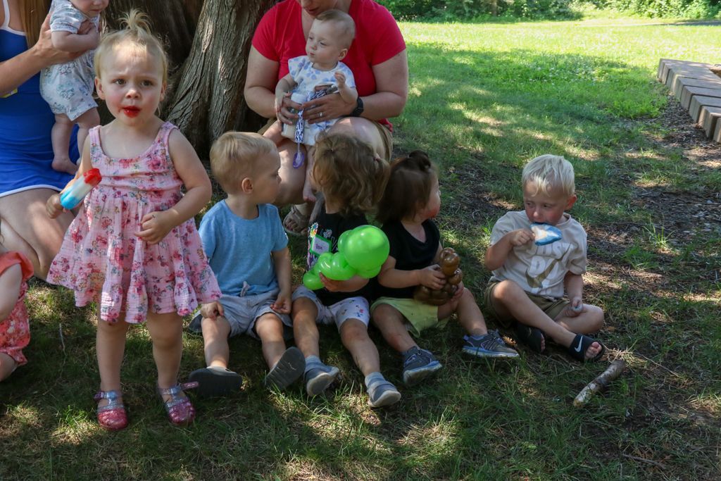 Children sitting on grass under a tree, several holding toys, with adults nearby. Sunny day.