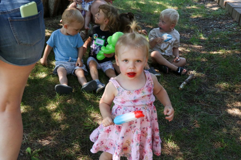 A young child in a pink dress holding a popsicle, surrounded by other children sitting on grass.