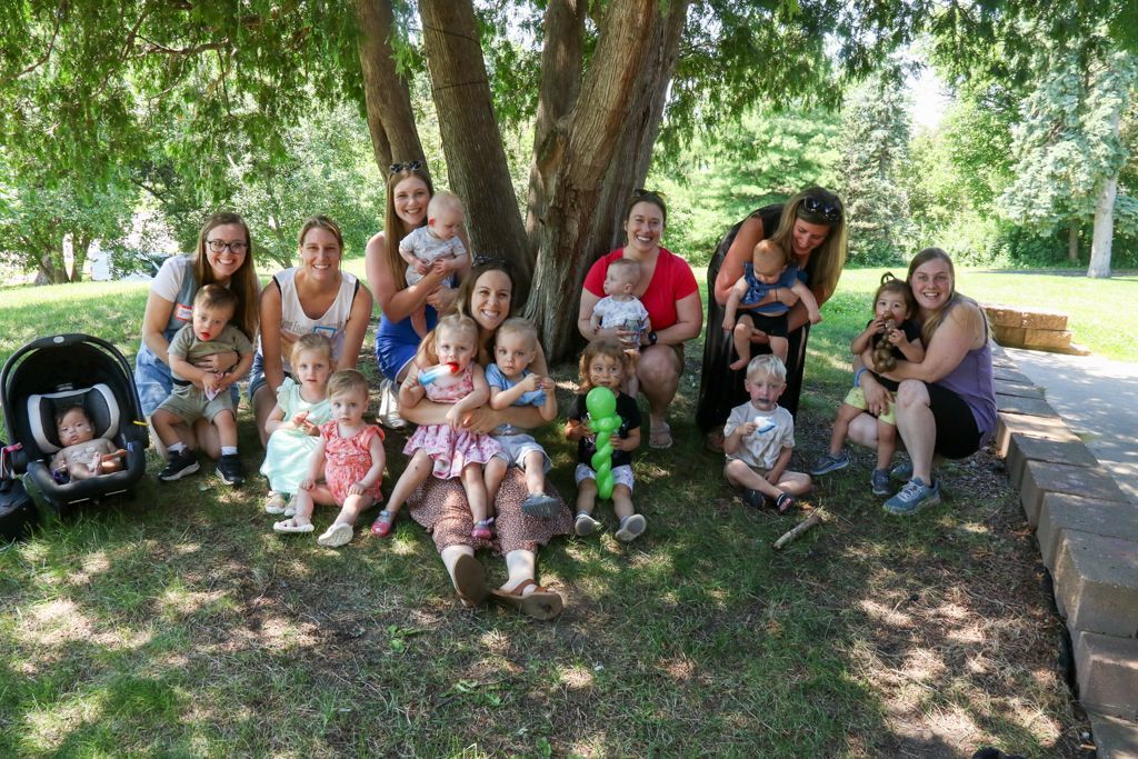 Group of adults and babies under a tree, posing for a photo outdoors on a sunny day.