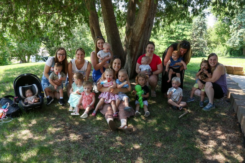 Group of adults and toddlers under a tree, smiling. Some hold babies; others sit on the grass. Sunny outdoor setting.