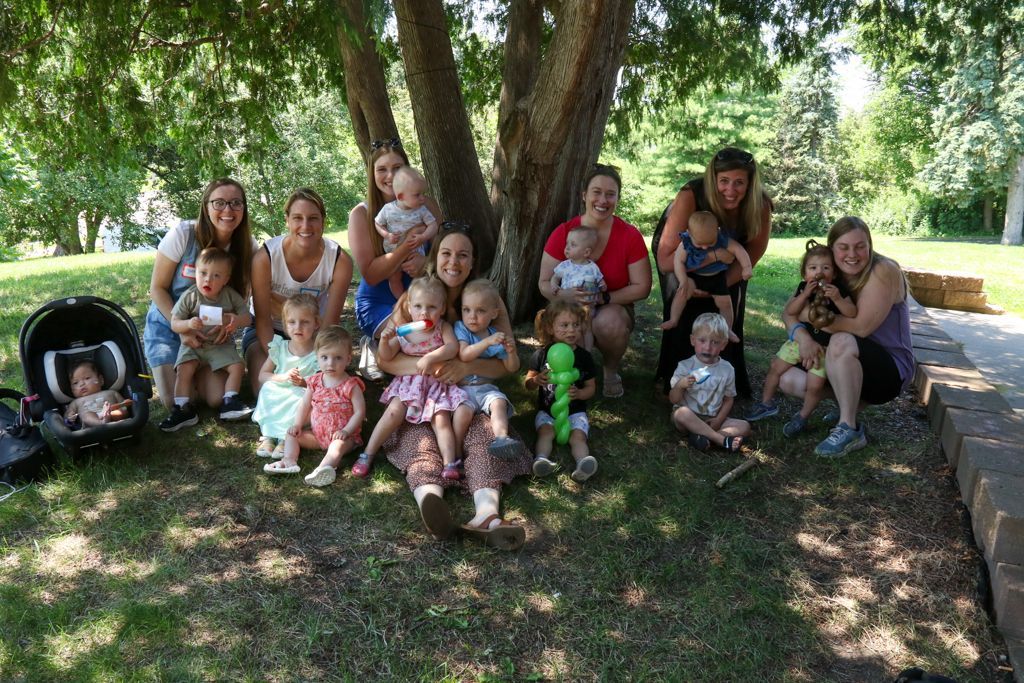 Group of adults with toddlers sit beneath a tree on grass.
