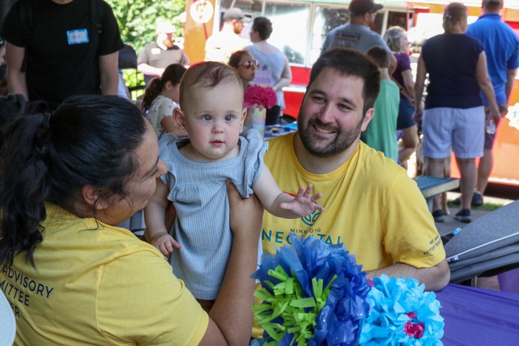 Woman holding baby, smiling man, and colorful decorations at an outdoor event.