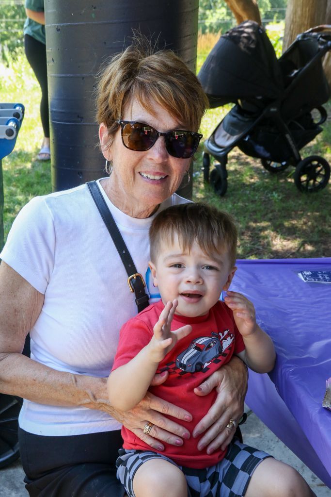 Woman in sunglasses holds a toddler in a red shirt outdoors. Both smile; the child gestures.