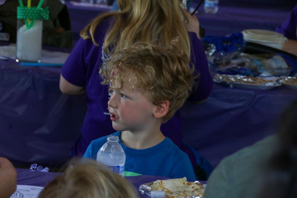 Boy with curly hair looking to the side, seated at a purple-covered table.
