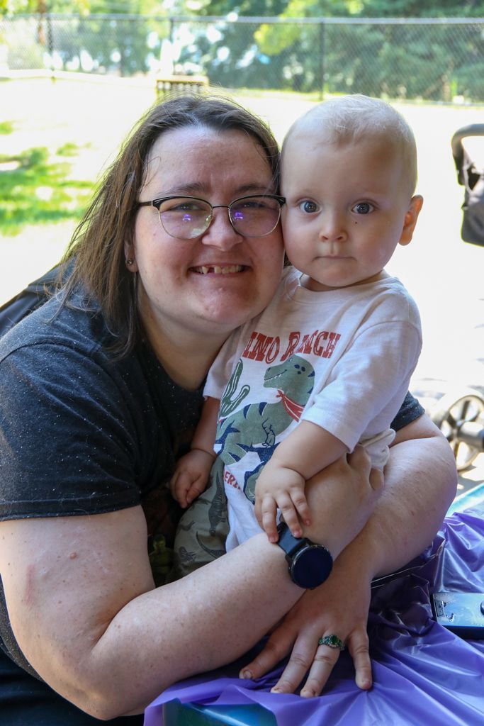 Woman in glasses holding a baby wearing a dinosaur t-shirt, both smiling outdoors.
