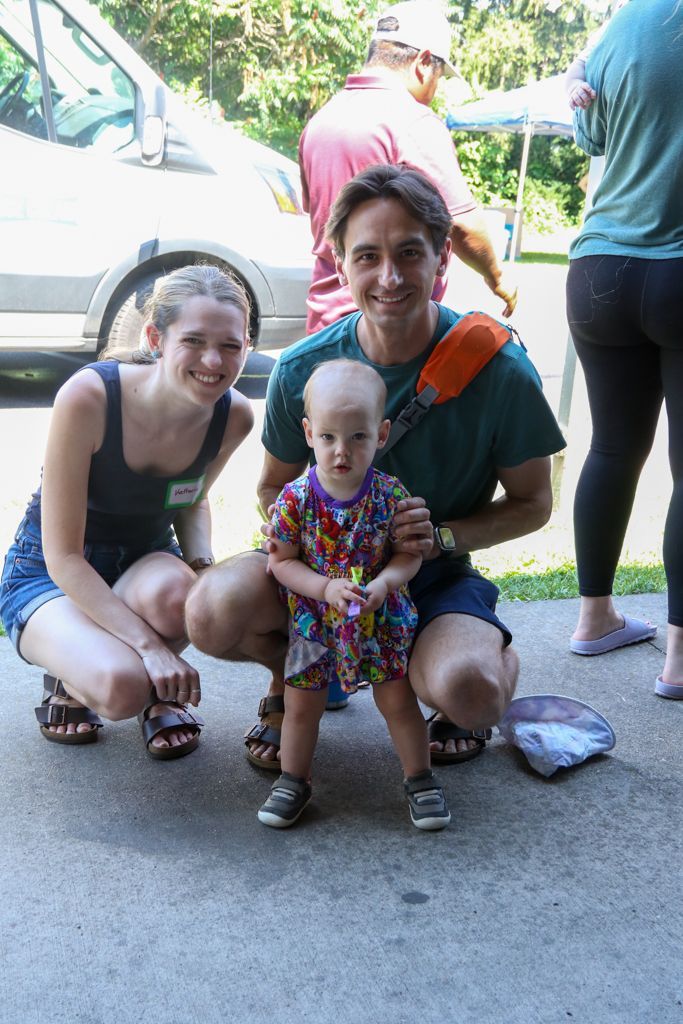 Family smiling outdoors: two adults squatting, child standing between them, sunny day.